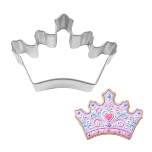 May include: A silver-colored metal crown-shaped cookie cutter next to a decorated crown-shaped cookie. The cookie is decorated with pink, purple, blue, and gold icing, featuring a heart and ornate details. The cookie cutter is made of shiny metal.