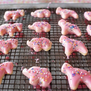 May include: Pink elephant-shaped cookies with a glossy glaze and colorful sprinkles rest on a wire rack. The cookies are arranged on a metal baking sheet, showcasing a delightful treat.