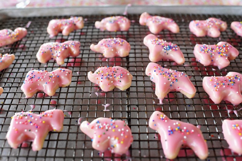 May include: Pink elephant-shaped cookies with a glossy glaze and colorful sprinkles rest on a wire rack. The cookies are arranged on a metal baking sheet, showcasing a delightful treat.
