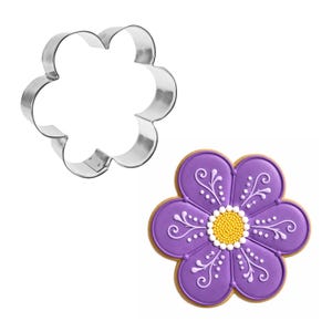May include: A silver-colored metal flower-shaped cookie cutter and a matching flower-shaped sugar cookie decorated with purple icing, white piped details, and a yellow beaded center. The cookie has a light brown baked edge.