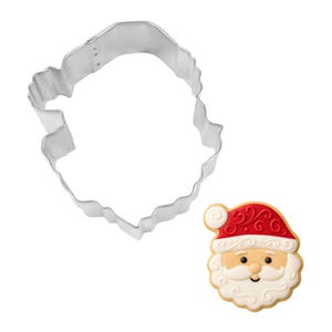 May include: A stainless steel Santa Claus face cookie cutter next to a decorated Santa Claus cookie. The cookie has a red hat with white trim, a white beard, and a smiling face. The cookie cutter is in the shape of Santa's face.