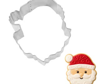 May include: A stainless steel Santa Claus face cookie cutter next to a decorated Santa Claus cookie. The cookie has a red hat with white trim, a white beard, and a smiling face. The cookie cutter is in the shape of Santa's face.