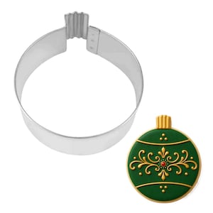 May include: A silver metal cookie cutter in the shape of a Christmas ornament, next to a decorated green cookie. The cookie has gold filigree and a red center, with a gold top. The cookie cutter is round with a ribbed top.