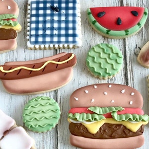 May include: Assortment of decorated sugar cookies in the shape of a hamburger, hot dog, watermelon slice, and a picnic blanket with an ant. The cookies are decorated with icing in various colors.