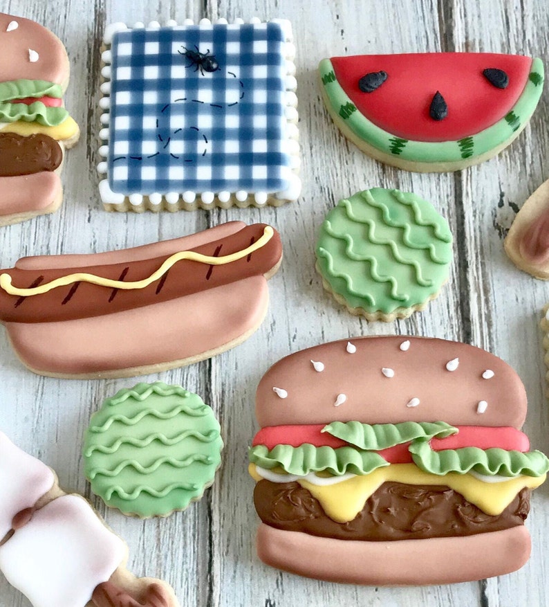 May include: Assortment of decorated sugar cookies in the shape of a hamburger, hot dog, watermelon slice, and a picnic blanket with an ant. The cookies are decorated with icing in various colors.