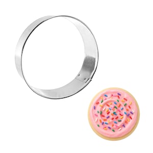 May include: A stainless steel circular cookie cutter next to a frosted sugar cookie with colorful sprinkles. The cookie has a pink frosting swirl and a light brown cookie base. The cutter is a ring shape.