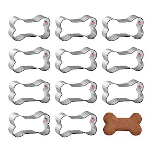 May include: A collection of twelve stainless steel dog bone-shaped cookie cutters, with one baked dog biscuit. The cookie cutters are arranged in three rows of four. Each cutter has a small American flag emblem.
