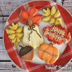May include: A plate of decorated Thanksgiving cookies. The cookies are shaped like fall leaves, pumpkins, and acorns. One cookie says "Happy Thanksgiving". The plate has a red plaid pattern.
