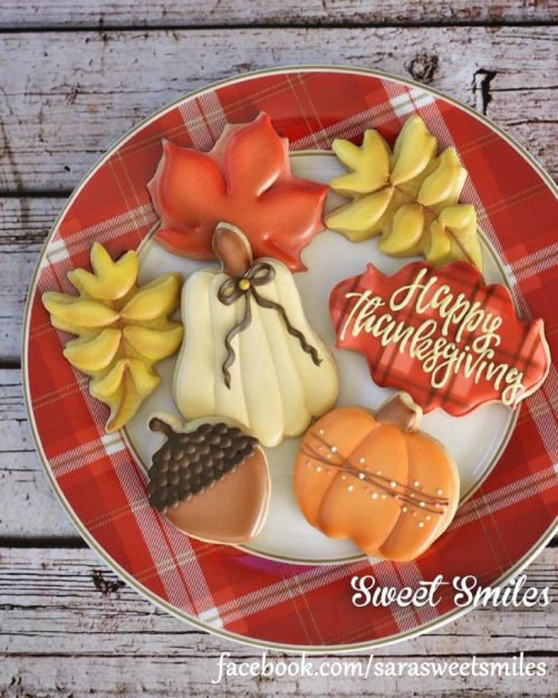 May include: A plate of decorated Thanksgiving cookies. The cookies are shaped like fall leaves, pumpkins, and acorns. One cookie says "Happy Thanksgiving". The plate has a red plaid pattern.