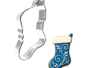May include: A stainless steel stocking-shaped cookie cutter next to a decorated stocking cookie. The cookie is blue with white floral designs and a white cuff. The cookie has a light brown border.