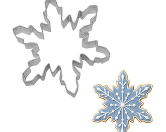 May include: A stainless steel snowflake-shaped cookie cutter next to a decorated snowflake cookie. The cookie has a light blue background with white icing details and a golden-brown edge. The cutter is a simple outline.