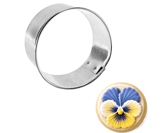 May include: A stainless steel round cookie cutter next to a decorated cookie. The cookie is round with a yellow, white, and blue pansy design. The cookie cutter is made of shiny, silver metal.