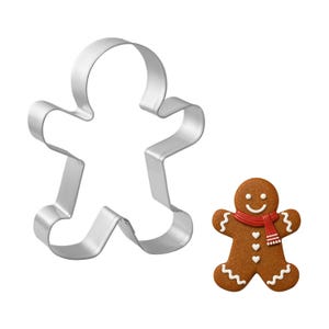 May include: A silver metal gingerbread man cookie cutter next to a decorated gingerbread man cookie. The cookie has a red scarf, white icing details, and a smiling face. The cutter is in the shape of a person with arms and legs.