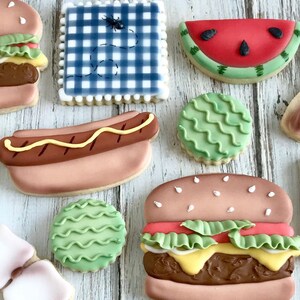 May include: Assortment of decorated sugar cookies in the shape of summer picnic foods. Includes a hamburger, watermelon slice, hot dog, and a blue gingham square with a bug design. The cookies are on a light wood surface.