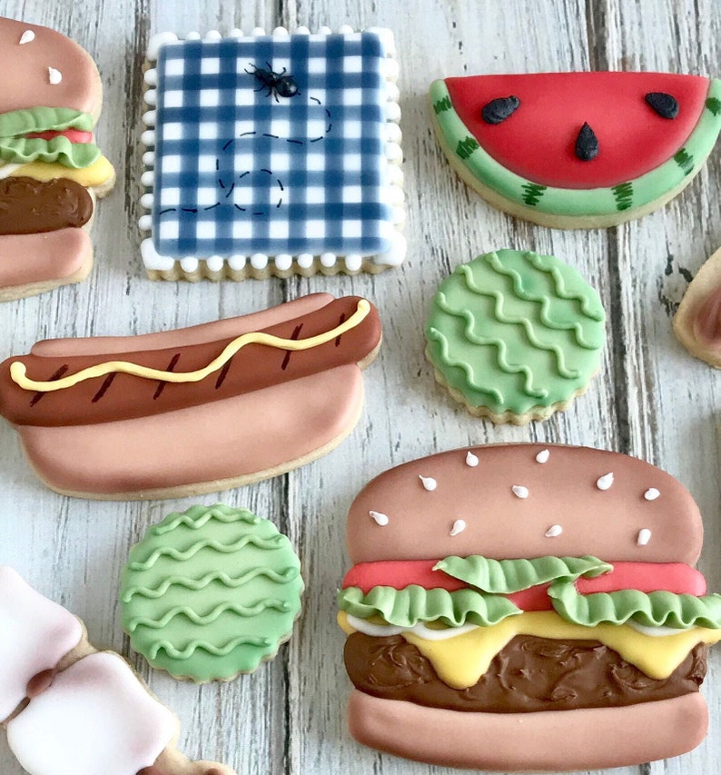 May include: Assortment of decorated sugar cookies in the shape of summer picnic foods. Includes a hamburger, watermelon slice, hot dog, and a blue gingham square with a bug design. The cookies are on a light wood surface.