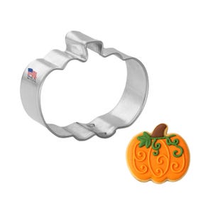 May include: A silver pumpkin-shaped cookie cutter next to a decorated pumpkin cookie. The cookie is orange with brown and green details. The cookie cutter has the text "Made in the USA" and a small American flag.