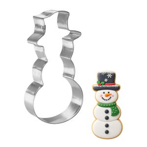 May include: A snowman-shaped cookie cutter next to a decorated snowman cookie. The cookie cutter is made of shiny, silver metal. The cookie is decorated with a black top hat, green scarf, and black buttons. The snowman has a carrot nose and a smiling face.
