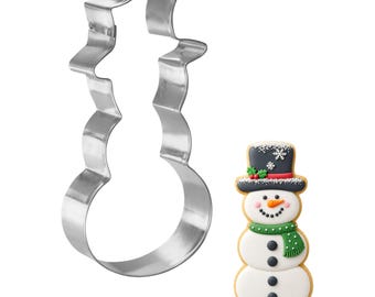 May include: A snowman-shaped cookie cutter next to a decorated snowman cookie. The cookie cutter is made of shiny, silver metal. The cookie is decorated with a black top hat, green scarf, and black buttons. The snowman has a carrot nose and a smiling face.