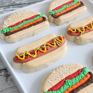 May include: An assortment of decorated sugar cookies shaped like hamburgers and hot dogs. The cookies feature colorful frosting details, including red, green, yellow, and brown, on a light-colored cookie base. Some cookies have sesame seeds.