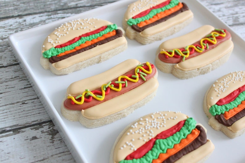 May include: An assortment of decorated sugar cookies shaped like hamburgers and hot dogs. The cookies feature colorful frosting details, including red, green, yellow, and brown, on a light-colored cookie base. Some cookies have sesame seeds.
