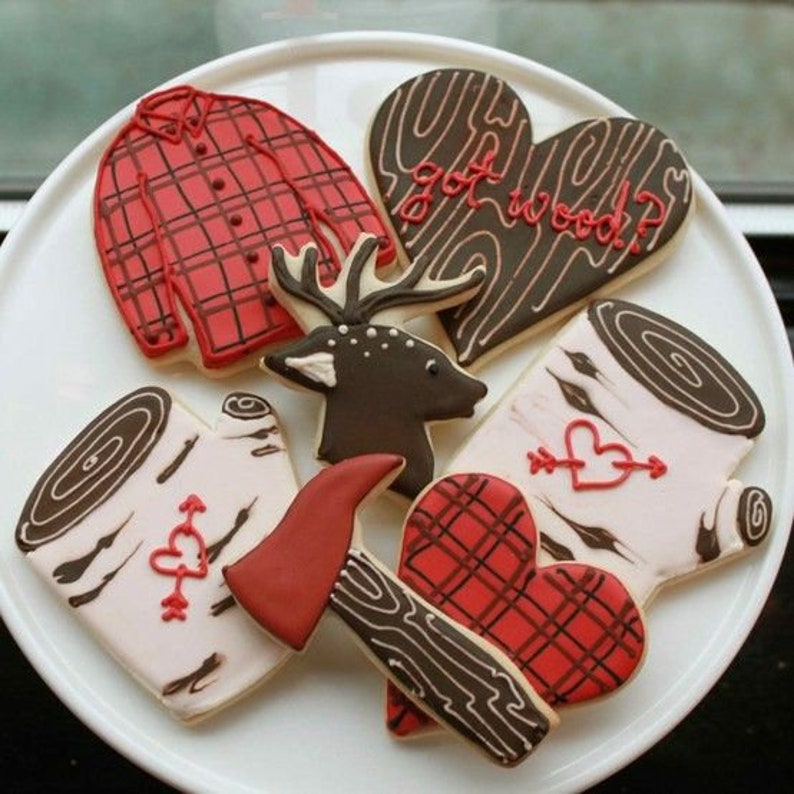 May include: A selection of decorated biscuits in various shapes, including a red plaid shirt, a deer head, a red axe, and heart-shaped biscuits with wood grain and the text "got wood?". The biscuits are arranged on a white plate.