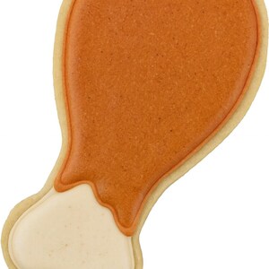 May include: A decorated cookie in the shape of a turkey leg. The cookie has a light tan border, with a textured orange-brown top and a cream-colored bottom, resembling the meat and bone of a cooked turkey leg.