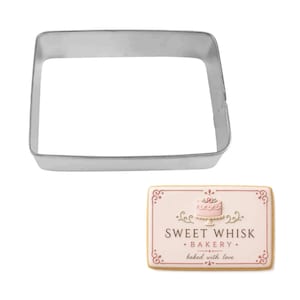 May include: A square-shaped, silver-colored cookie cutter next to a rectangular cookie. The cookie has a pink background with the text "SWEET WHISK BAKERY" and a cake illustration. The cookie is decorated with pink and white details.