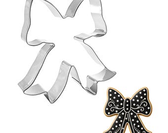 May include: A shiny, silver-colored metal bow-shaped cookie cutter next to a decorated bow-shaped cookie. The cookie is black with white polka dots and decorative swirls, outlined in gold.