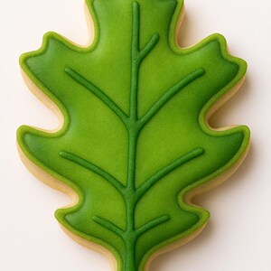 May include: A leaf-shaped cookie with a green, detailed leaf design. The cookie has a light green center with darker green veins and edges. The outer edge of the cookie is a light tan color, creating a contrast with the green leaf.