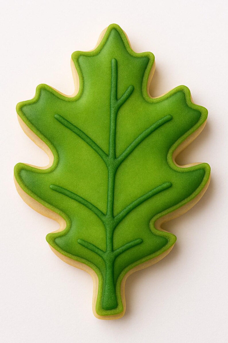 May include: A leaf-shaped cookie with a green, detailed leaf design. The cookie has a light green center with darker green veins and edges. The outer edge of the cookie is a light tan color, creating a contrast with the green leaf.