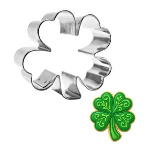May include: A shiny, silver metal shamrock cookie cutter next to a decorated shamrock cookie. The cookie is green with darker green and yellow details. The cookie cutter is in the shape of a four-leaf clover.