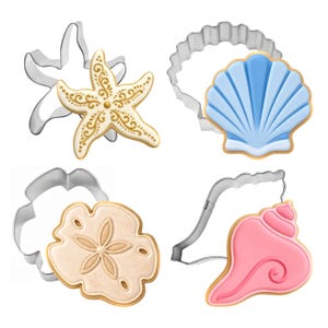 May include: A set of four metal cookie cutters paired with matching decorated cookies. The shapes include a gold-detailed starfish, a blue scalloped shell, a beige sand dollar, and a pink conch shell, each with its corresponding metal outline.
