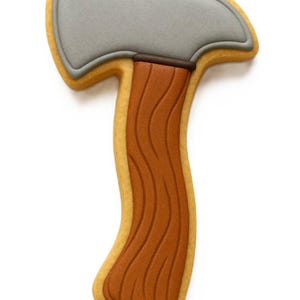 May include: An axe-shaped biscuit. The axe head is grey, the handle is brown with a wood grain effect, and the biscuit is outlined in yellow. The biscuit is set against a white backdrop.