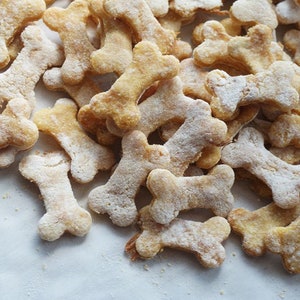 May include: A close-up of a pile of homemade dog treats shaped like bones. The treats are light brown and have a slightly powdery texture.