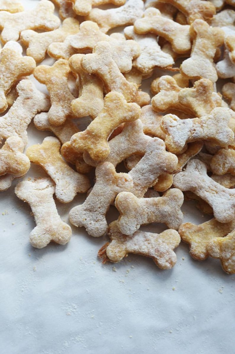 May include: A close-up of a pile of homemade dog treats shaped like bones. The treats are light brown and have a slightly powdery texture.
