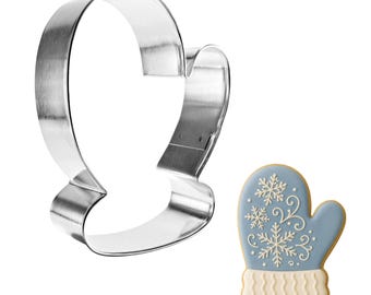May include: A stainless steel mitten-shaped cookie cutter next to a decorated mitten-shaped cookie. The cookie is light blue with white snowflake and swirl designs, and a cream-colored cuff and border. The cutter is for baking.