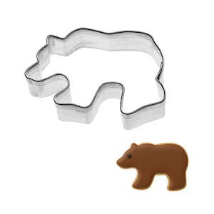 May include: A stainless steel bear-shaped cookie cutter next to a brown bear-shaped cookie. The cookie cutter is a simple outline of a bear. The cookie is brown with a darker brown eye.