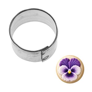 May include: A round, stainless steel cookie cutter next to a decorated cookie. The cookie is round with a purple and white pansy design and a yellow center. The cookie cutter is a cylinder shape.