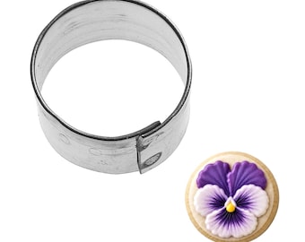 May include: A round, stainless steel cookie cutter next to a decorated cookie. The cookie is round with a purple and white pansy design and a yellow center. The cookie cutter is a cylinder shape.