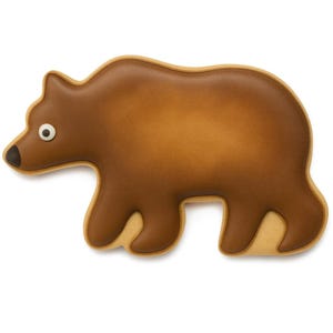 Op de afbeelding: Een bruine koek in de vorm van een beer met een lichtbruine omtrek en een donkerder bruin interieur. De beer heeft een kleine zwarte neus en een wit en zwart oog. De koek ligt op een witte achtergrond.