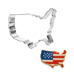 May include: A shiny, silver metal cookie cutter in the shape of the United States, next to a decorated cookie. The cookie is shaped like the U.S. with a red and white striped design and a blue section with white stars, resembling the American flag.