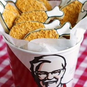 May include: A bucket of fried chicken cookies. The cookies are shaped like chicken drumsticks with a white bone and a crispy, golden-brown coating. The bucket is red and white, featuring a portrait of a man with glasses.