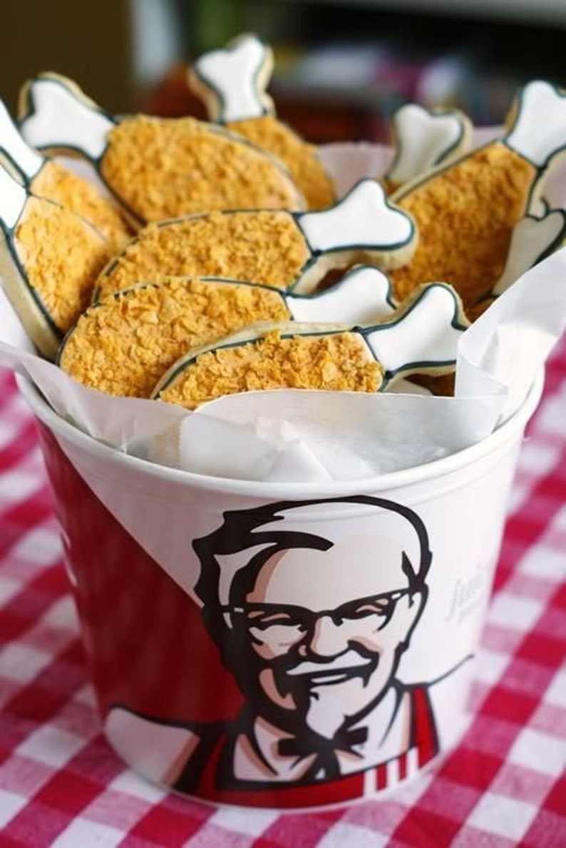 May include: A bucket of fried chicken cookies. The cookies are shaped like chicken drumsticks with a white bone and a crispy, golden-brown coating. The bucket is red and white, featuring a portrait of a man with glasses.