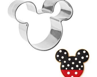 May include: A stainless steel Mickey Mouse-shaped cookie cutter next to a decorated cookie. The cookie has a black top with white polka dots and a red bottom, resembling Mickey's shorts.