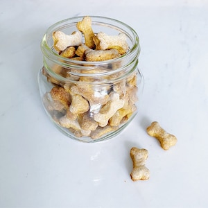 May include: A glass jar filled with dog biscuits in the shape of bones. The biscuits are a light brown color and have a slightly powdery texture. There are two additional biscuits on the white surface next to the jar.