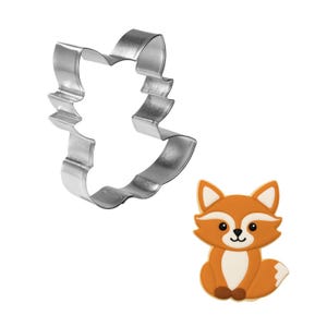 May include: A stainless steel fox-shaped cookie cutter next to a decorated fox cookie. The cookie is orange, white, and brown, with a smiling face. The cookie cutter is a simple outline of the fox shape.