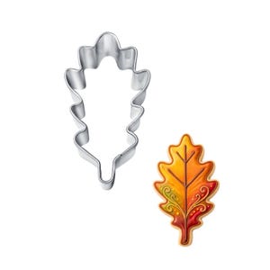May include: A stainless steel oak leaf-shaped cookie cutter next to a decorated cookie. The cookie is iced with a gradient of yellow, orange, and red, with intricate green and red decorative details.