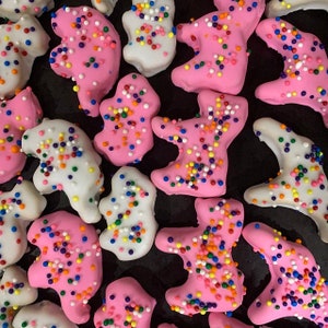 May include: Close-up of frosted cookies in the shape of teddy bears. Some are pink, others are white, and all are covered in colorful sprinkles. The cookies are arranged on a dark surface.