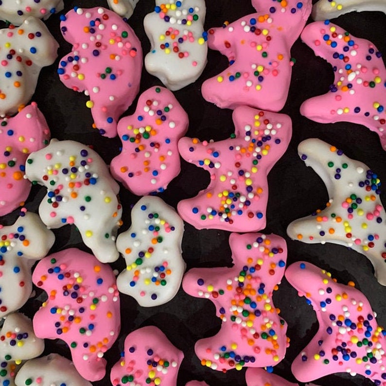 May include: Close-up of frosted cookies in the shape of teddy bears. Some are pink, others are white, and all are covered in colorful sprinkles. The cookies are arranged on a dark surface.