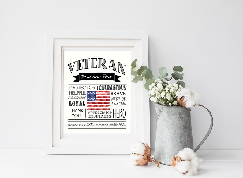 May include: A framed print with the word "VETERAN" at the top, featuring an American flag design. The print includes words like "Protector," "Courageous," and "Hero." A metal pitcher with flowers and cotton accents is to the right.